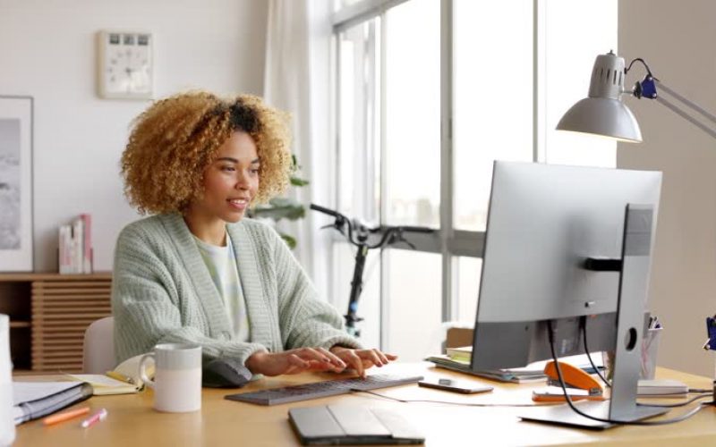 Confident businesswoman with Afro hairstyle using computer at desk. Female entrepreneur is working from home. She is telecommuting.